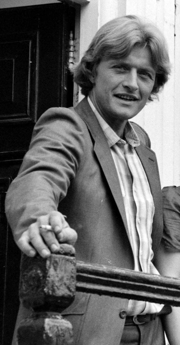 A young man with fair hair in a jacket leaning on a doorway rail