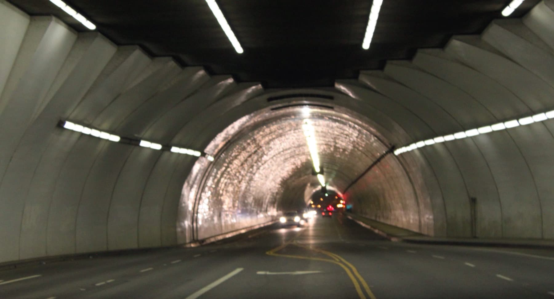 A curving white-tiled road tunnel lit by strip lights