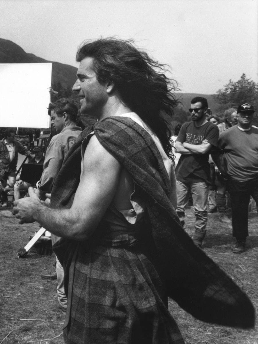 A long-haired man in a belted plaid stands side-on in the wind in front of film lights and crew