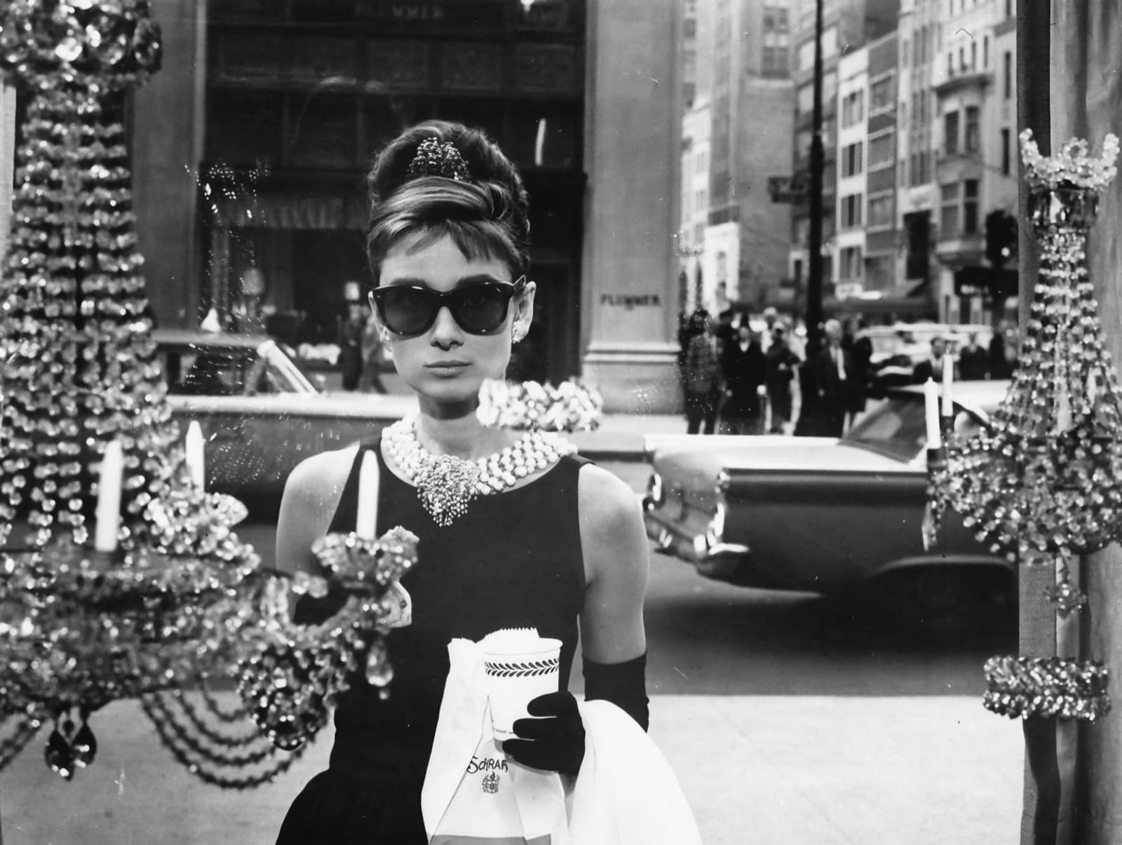 Audrey Hepburn in a black dress, dark glasses and pearls, holding a paper cup and a pastry bag, framed between crystal chandeliers in a shop window