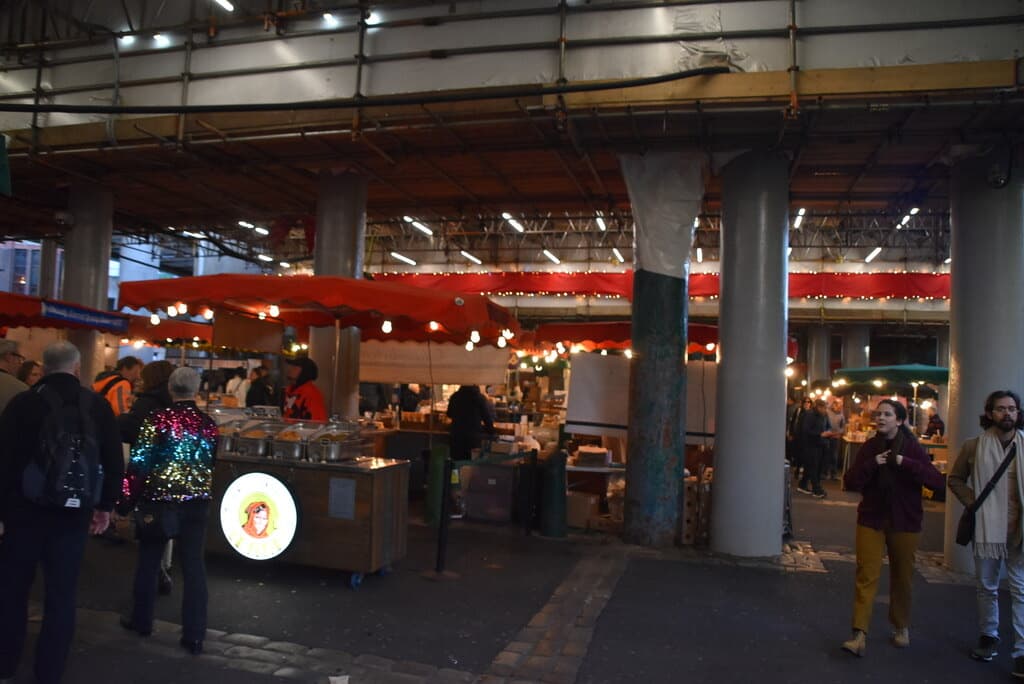 Market stalls under iron columns and red awnings, with shoppers walking between them