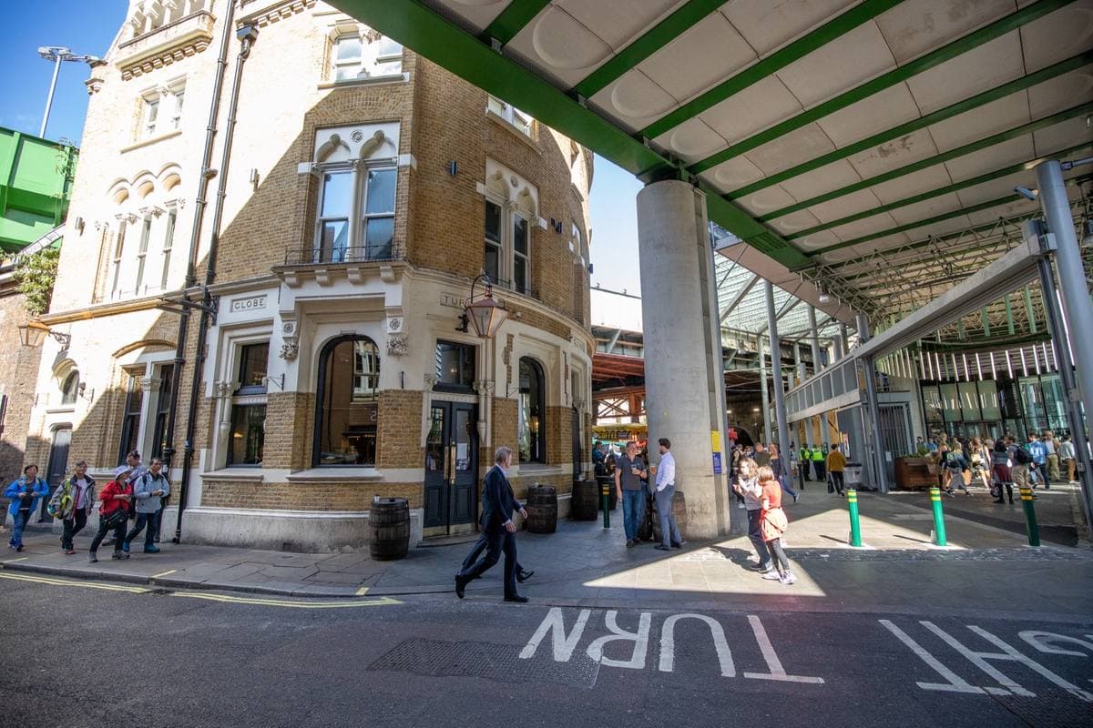 A corner pub under the green iron roof of a market, with people crossing the street in front