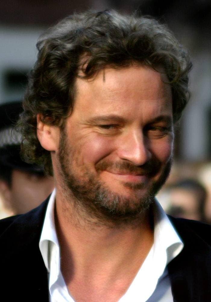Colin Firth with curly hair and stubble, smiling, in a dark jacket and open white shirt