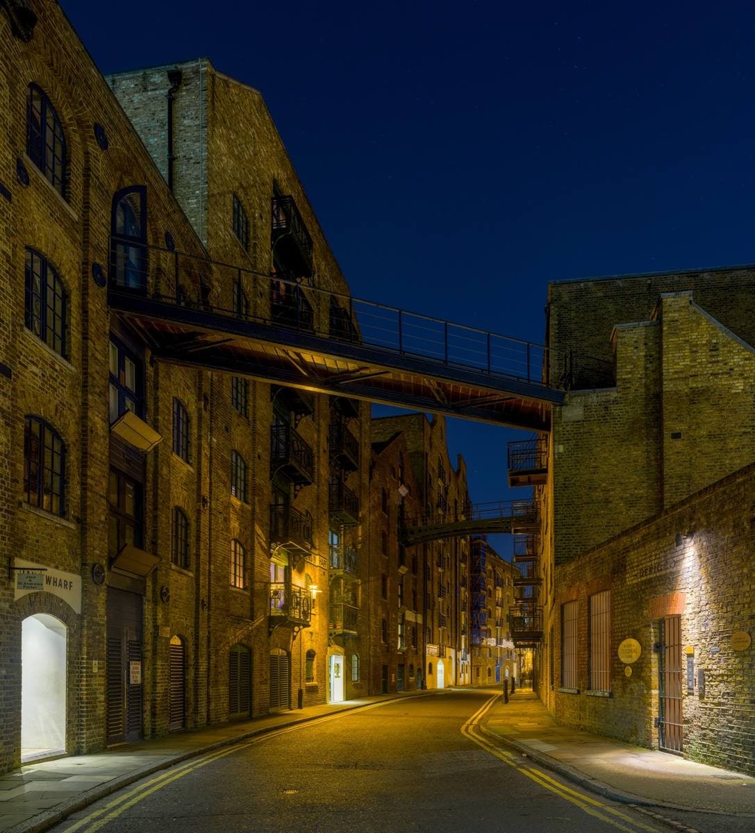 A narrow cobbled street between tall brick warehouses, crossed by iron walkways, lit gold under a dark blue sky
