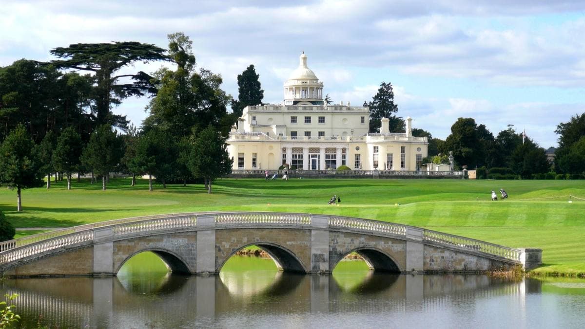 A white domed country house beyond a lake and a stone bridge on a golf course