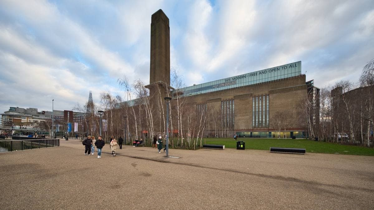 A huge brick former power station with a tall square chimney, behind a gravel square and birch trees