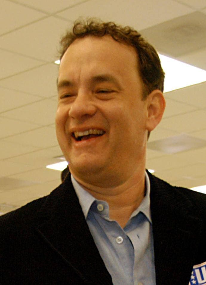 Tom Hanks laughing, in a dark jacket and open-necked blue shirt
