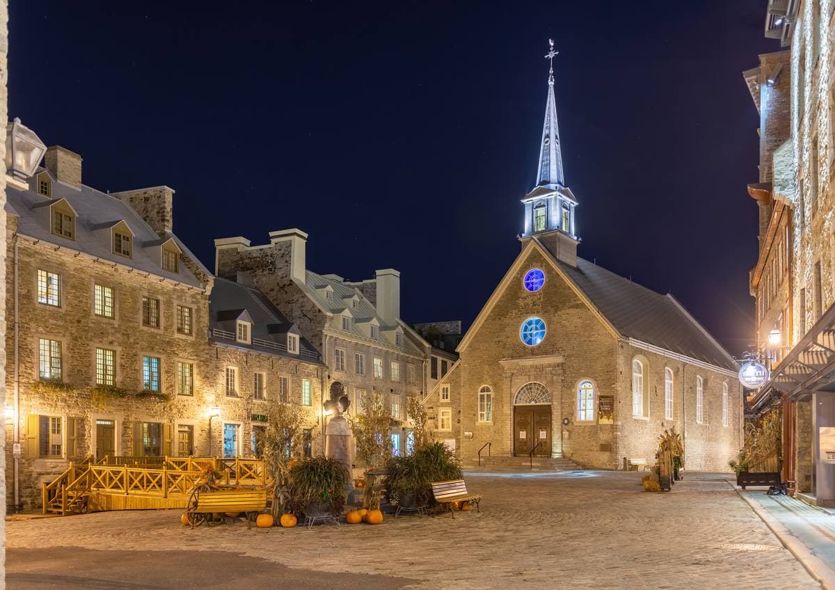 A small stone square at night, lit stone houses and a church with a pointed white spire