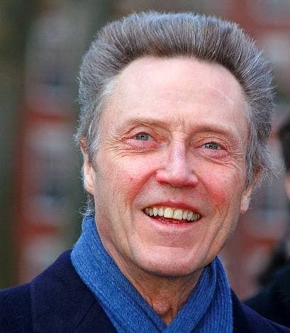 Christopher Walken smiling, his grey hair swept up, a blue scarf at his neck