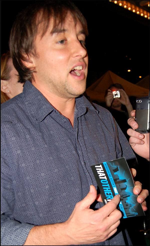 Richard Linklater mid-sentence, holding a newspaper, in a dark patterned shirt