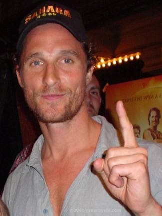 Matthew McConaughey in a baseball cap, smiling and holding up one finger