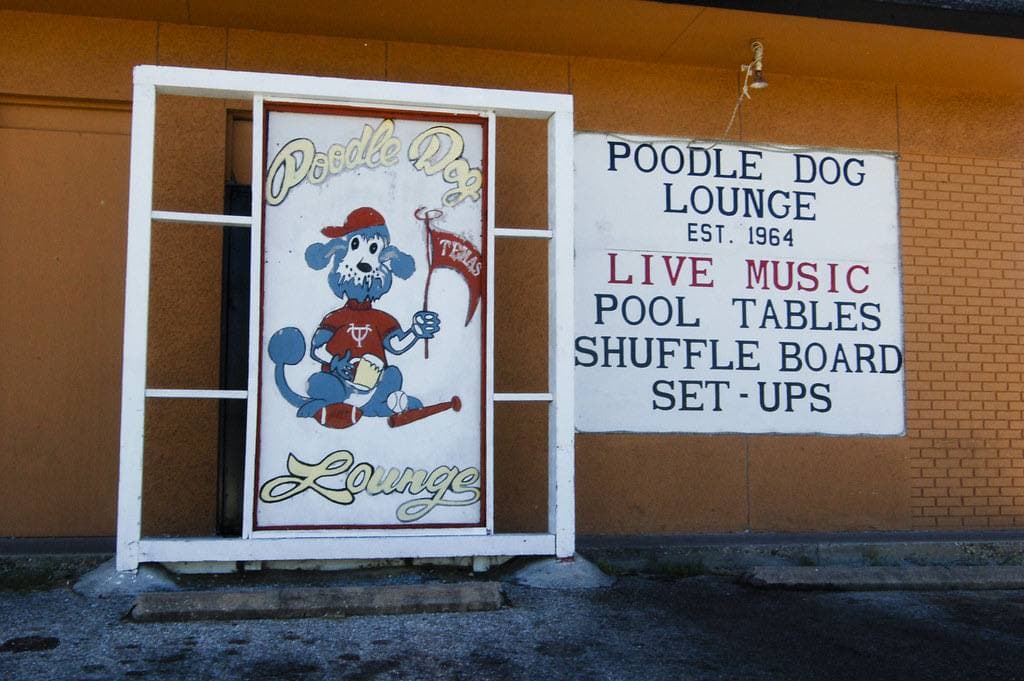 A painted Poodle Dog Lounge sign beside a board listing live music, pool tables and shuffle board
