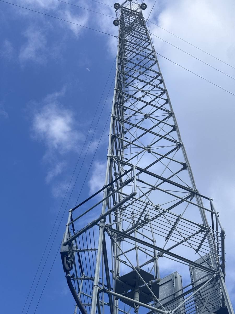 Looking up the steel lattice of a moonlight tower to its lamps, the moon in the blue sky beyond