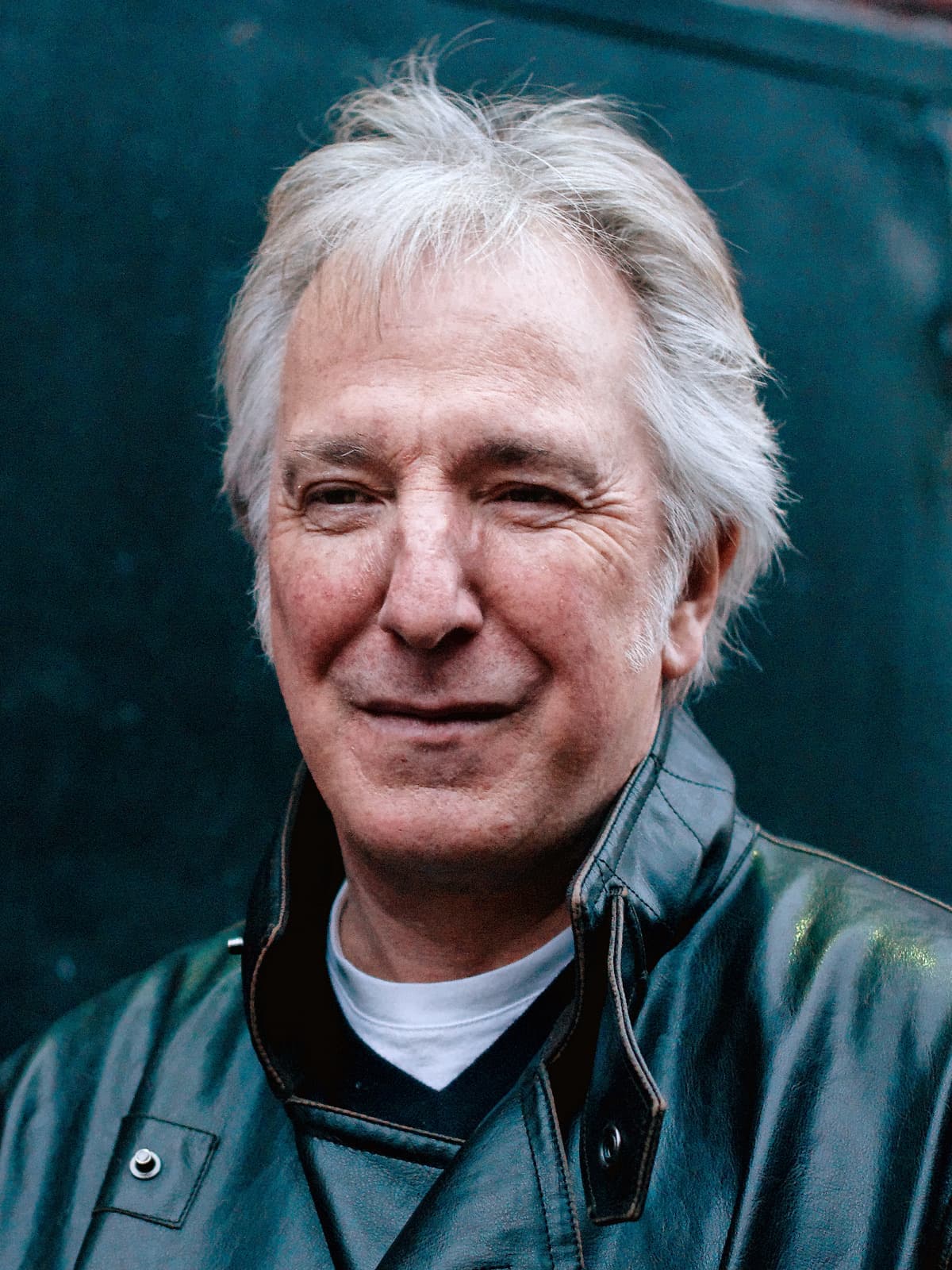 Alan Rickman in a leather jacket, smiling, against a dark green wall