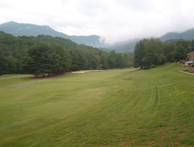 A green fairway lined with trees running towards misty mountains