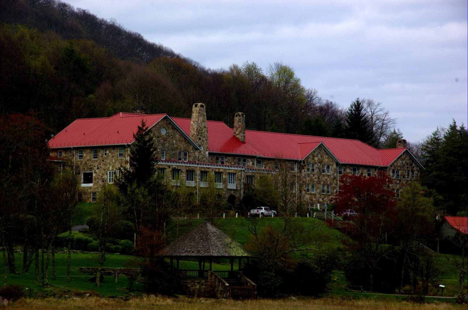 A long stone hotel with a red roof on a wooded slope, a small open gazebo on the lawn below