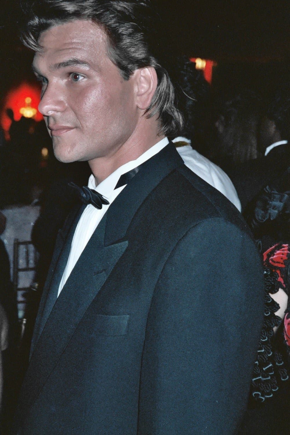 A man with swept-back hair in a black tuxedo and bow tie, seen in profile at night
