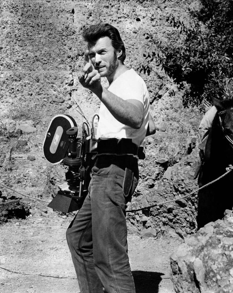 Clint Eastwood in a white T-shirt beside a film camera, pointing as he gives an instruction