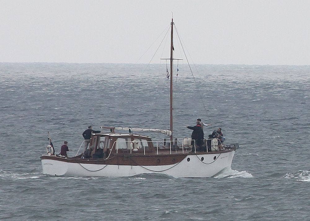 A varnished 1930s motor yacht with a white hull and a short mast, four figures on deck, on a choppy grey sea