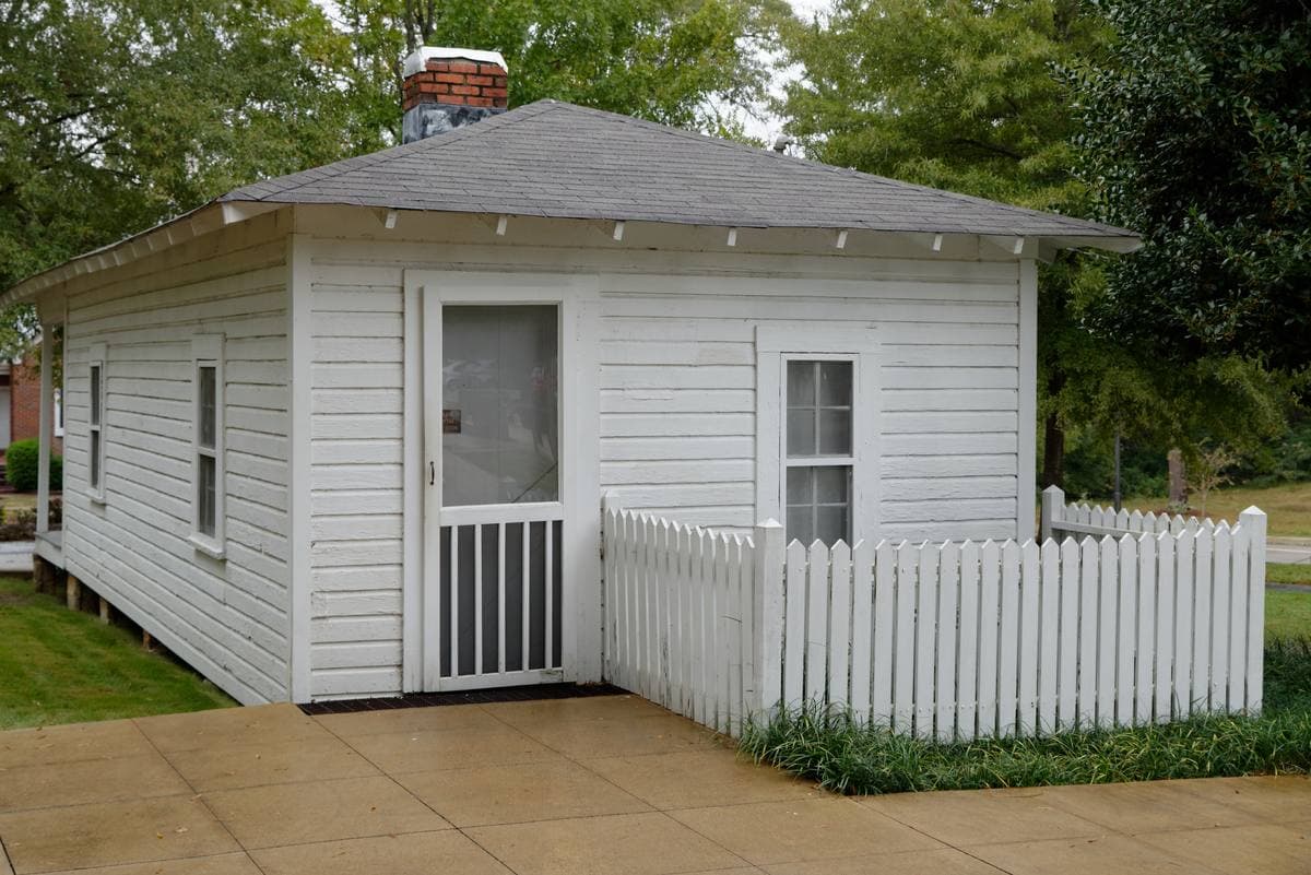 A small white shotgun house with a porch swing under trees