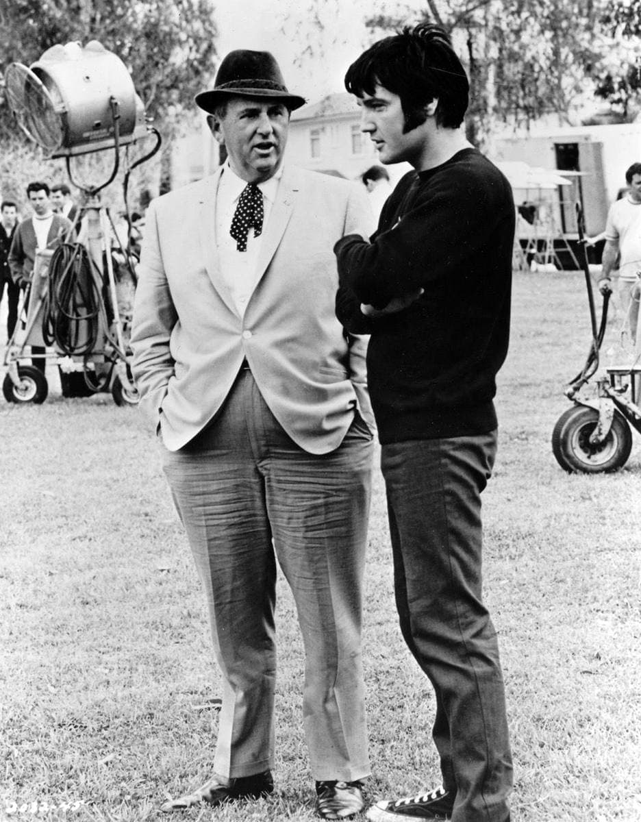 Colonel Tom Parker in a hat and pale jacket talking with Elvis Presley in a dark sweater outdoors