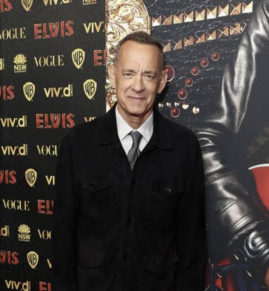 Tom Hanks in a dark jacket smiling in front of a printed backdrop