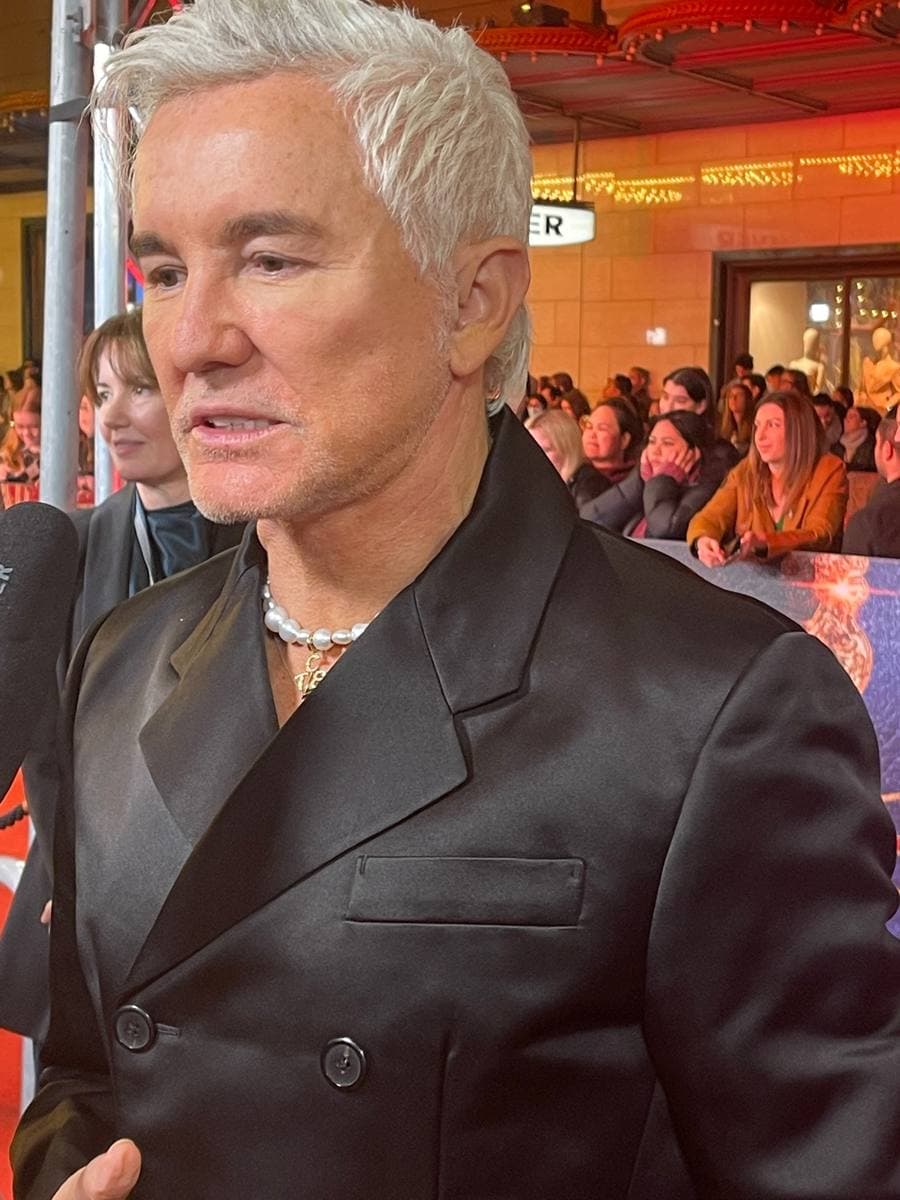 Baz Luhrmann with white hair and a pearl necklace in a black satin jacket