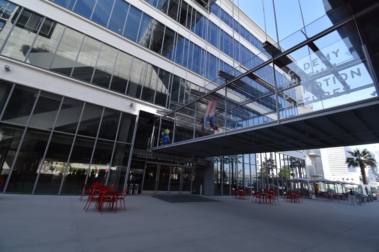 A glass-fronted museum building with a footbridge overhead and red chairs outside