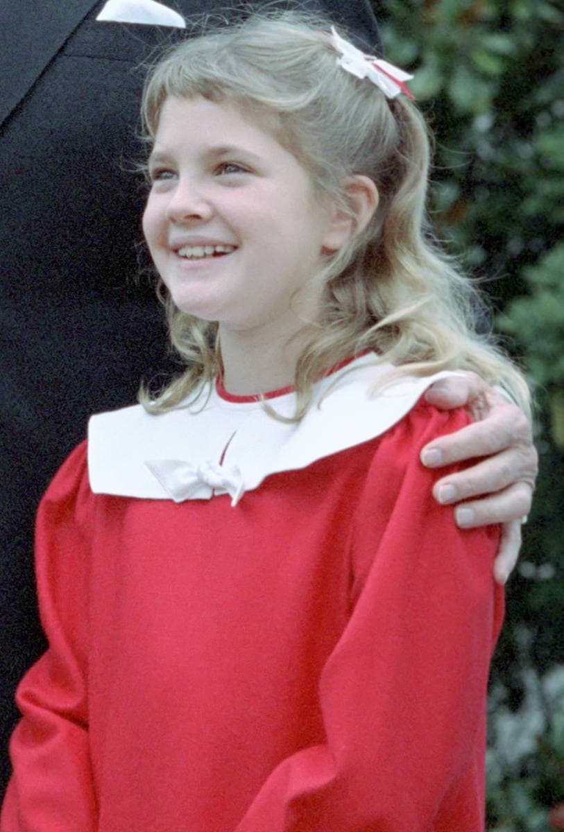 Drew Barrymore as a smiling girl in a red dress with a white collar and a bow in her hair