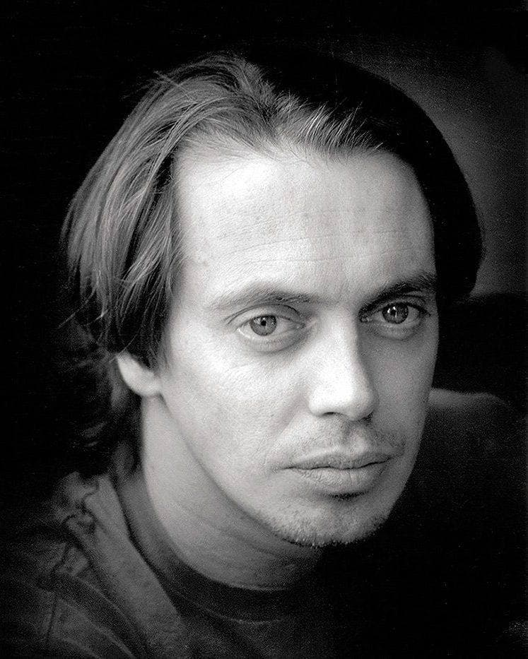 Black-and-white portrait of a young Steve Buscemi with long hair, looking into the camera