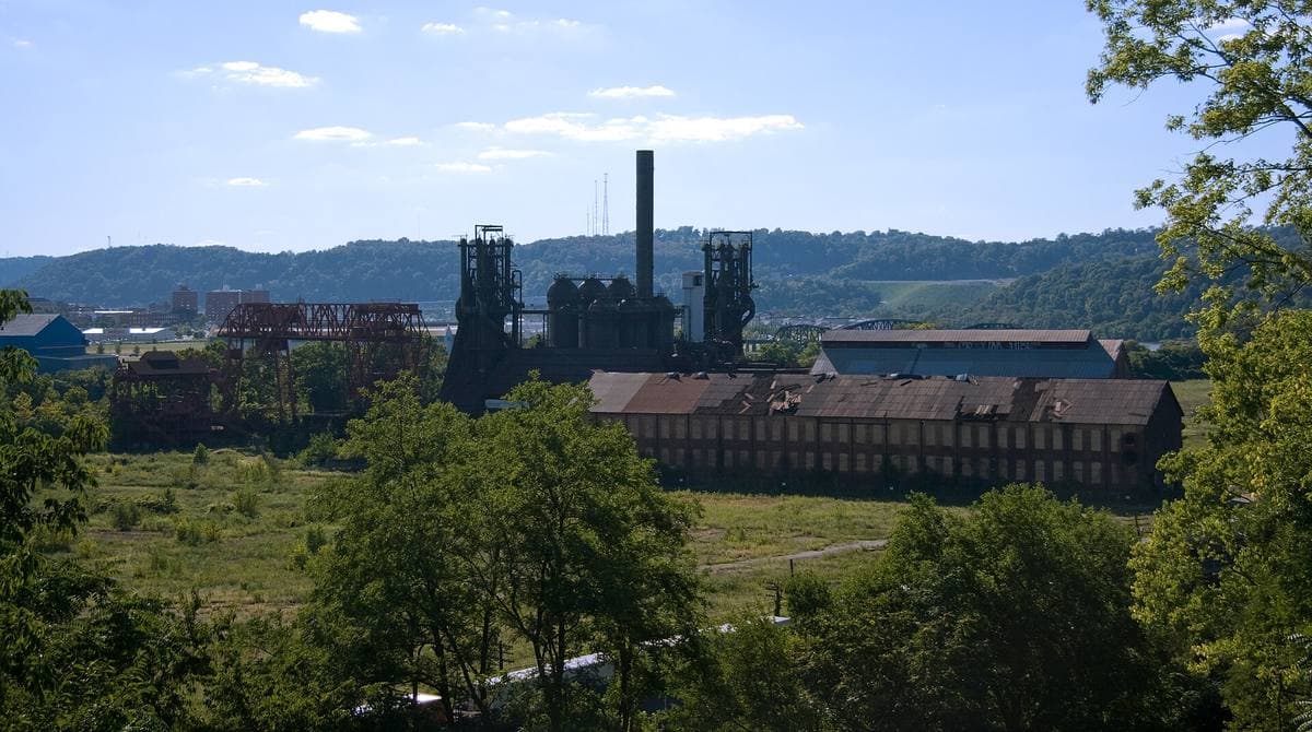 Two dark blast furnaces and a long brick building beyond a green river bank