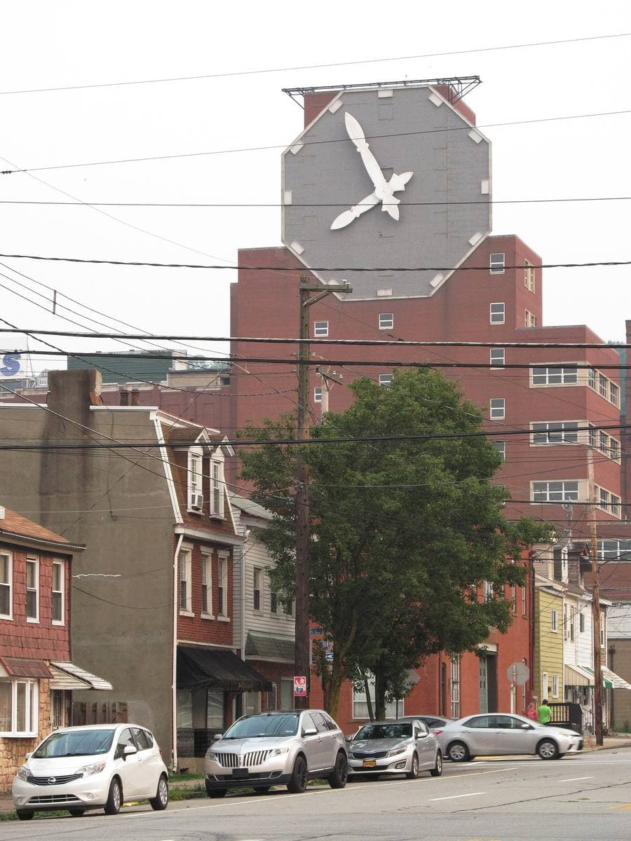 A huge square clock face on top of a brick brewery building, rowhouses and parked cars below