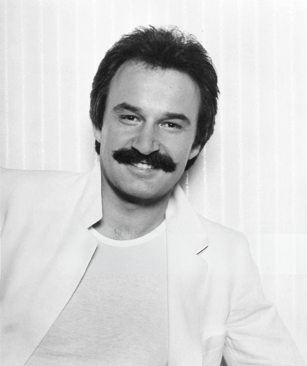 Giorgio Moroder, smiling under a thick moustache, in a white jacket and T-shirt
