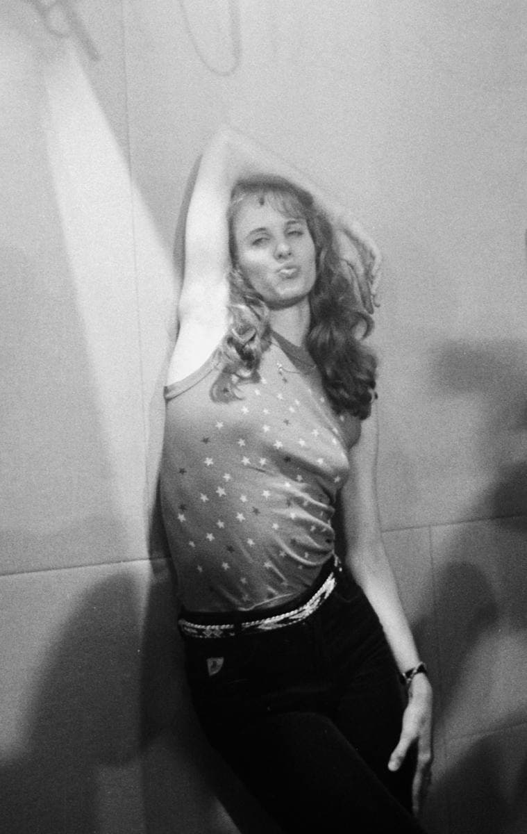 Lori Singer, young, with long wavy hair, one arm raised behind her head, leaning on a wall