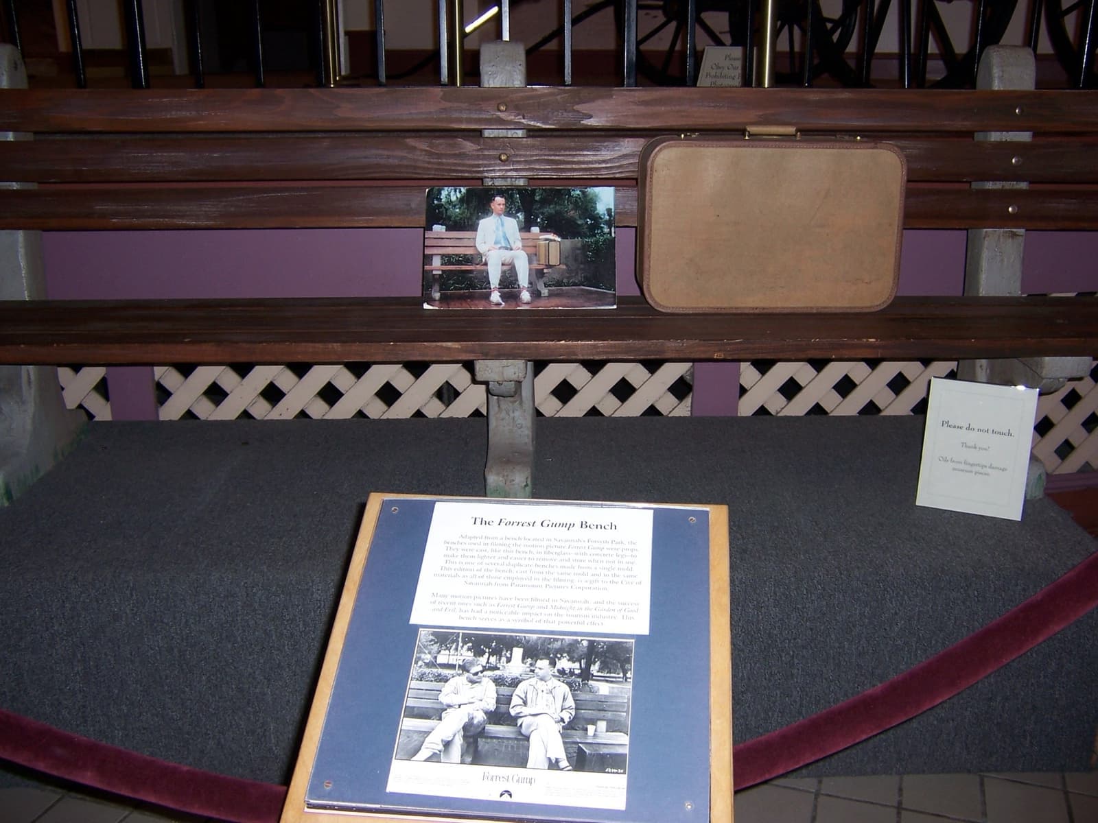 A long wooden-slatted bench behind a red rope with a brown suitcase on it and an information panel in front
