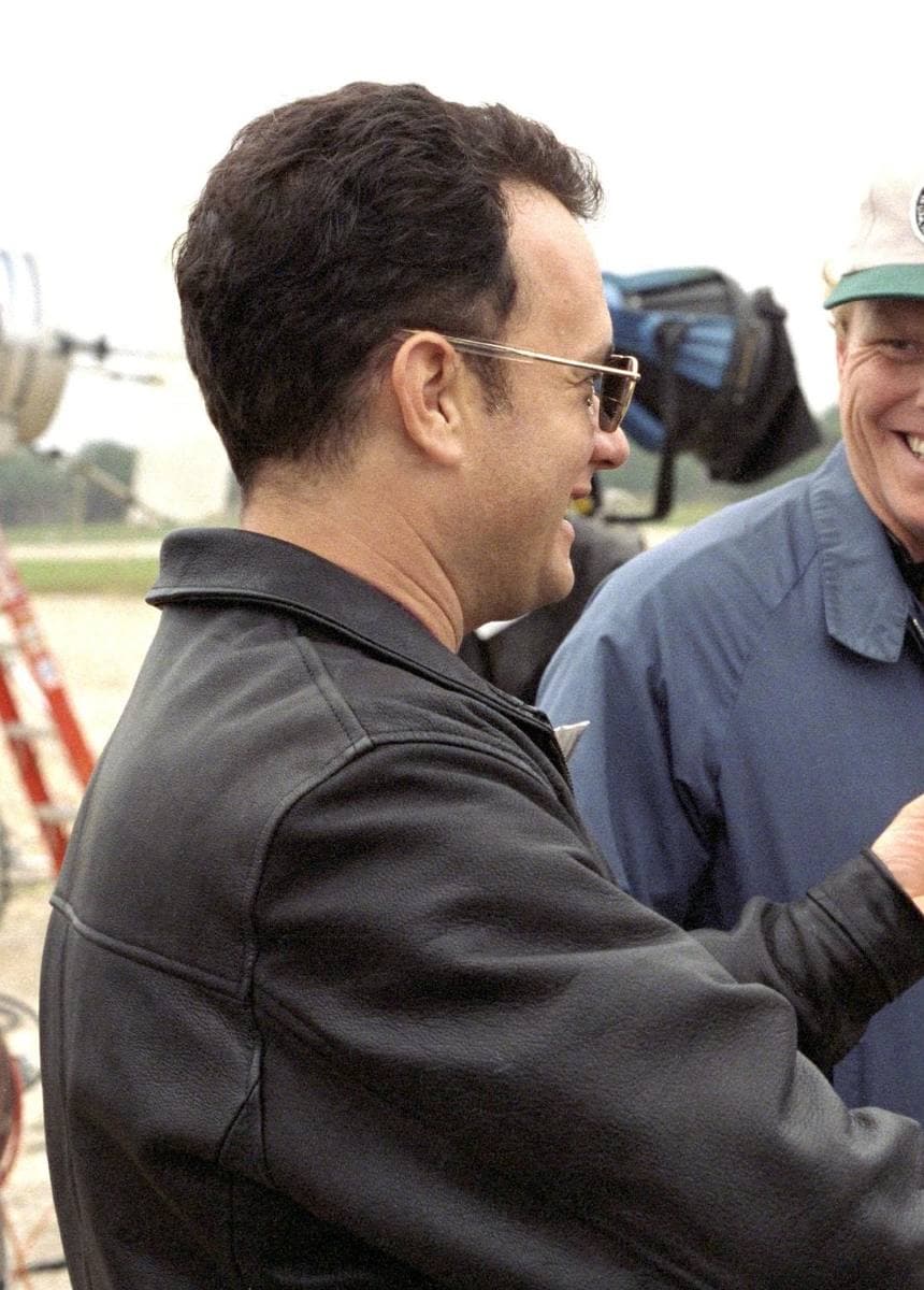 Tom Hanks in glasses and a black leather jacket, talking with a crew member outdoors