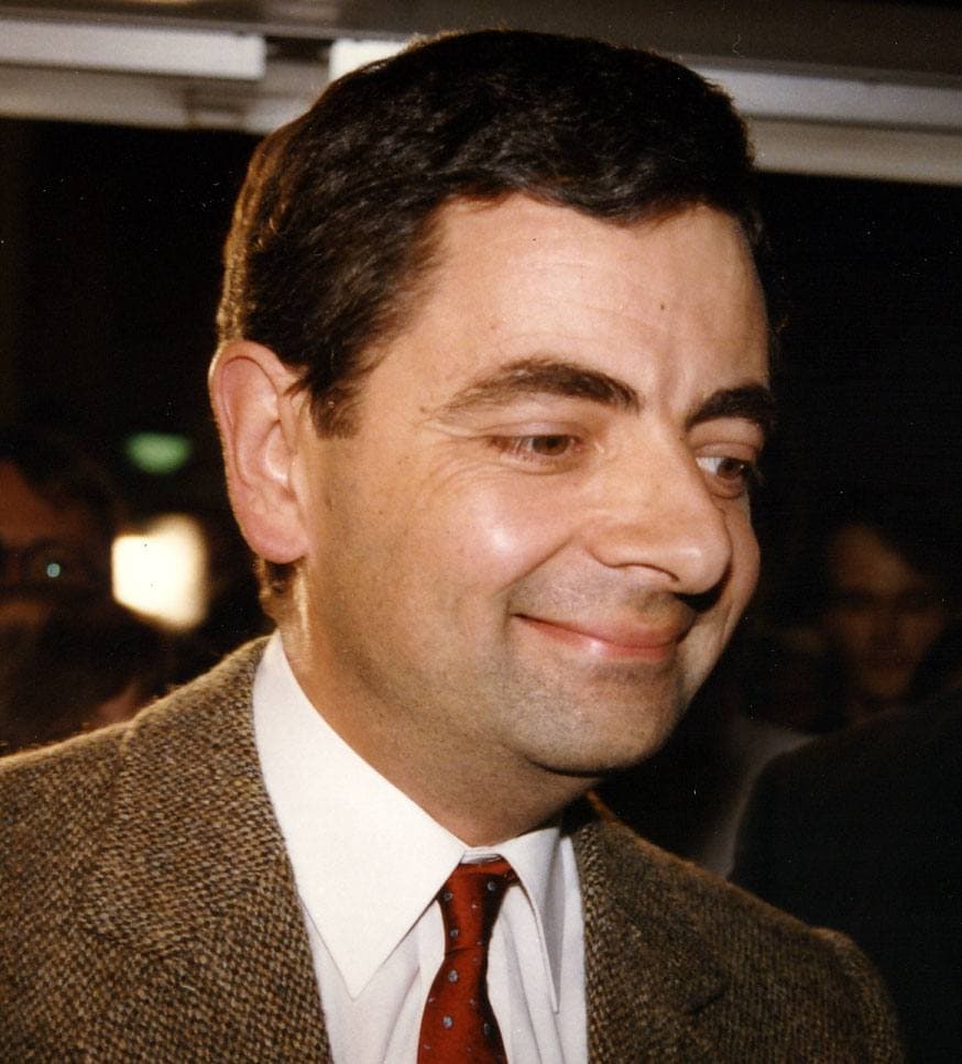 Rowan Atkinson in a tweed jacket and red tie, smiling sideways