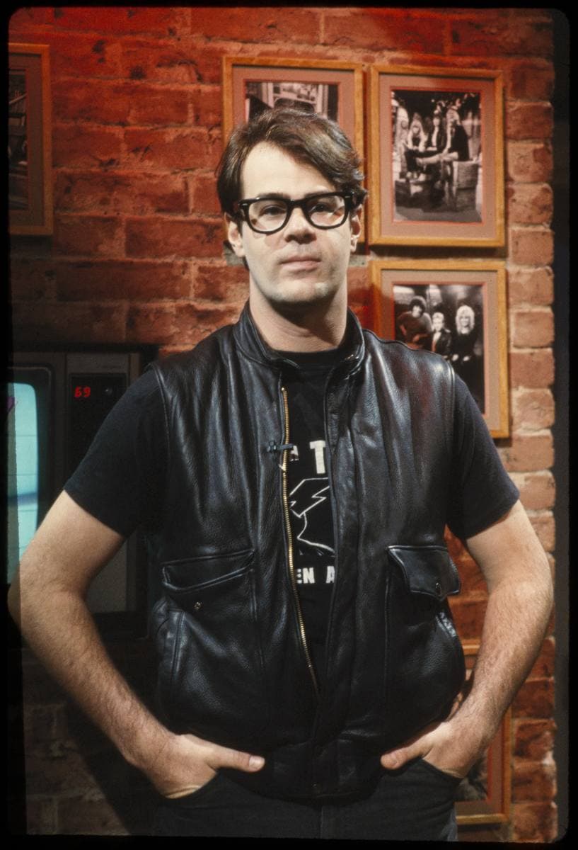 Dan Aykroyd in black-framed glasses and a leather waistcoat, hands in pockets, in front of a brick wall