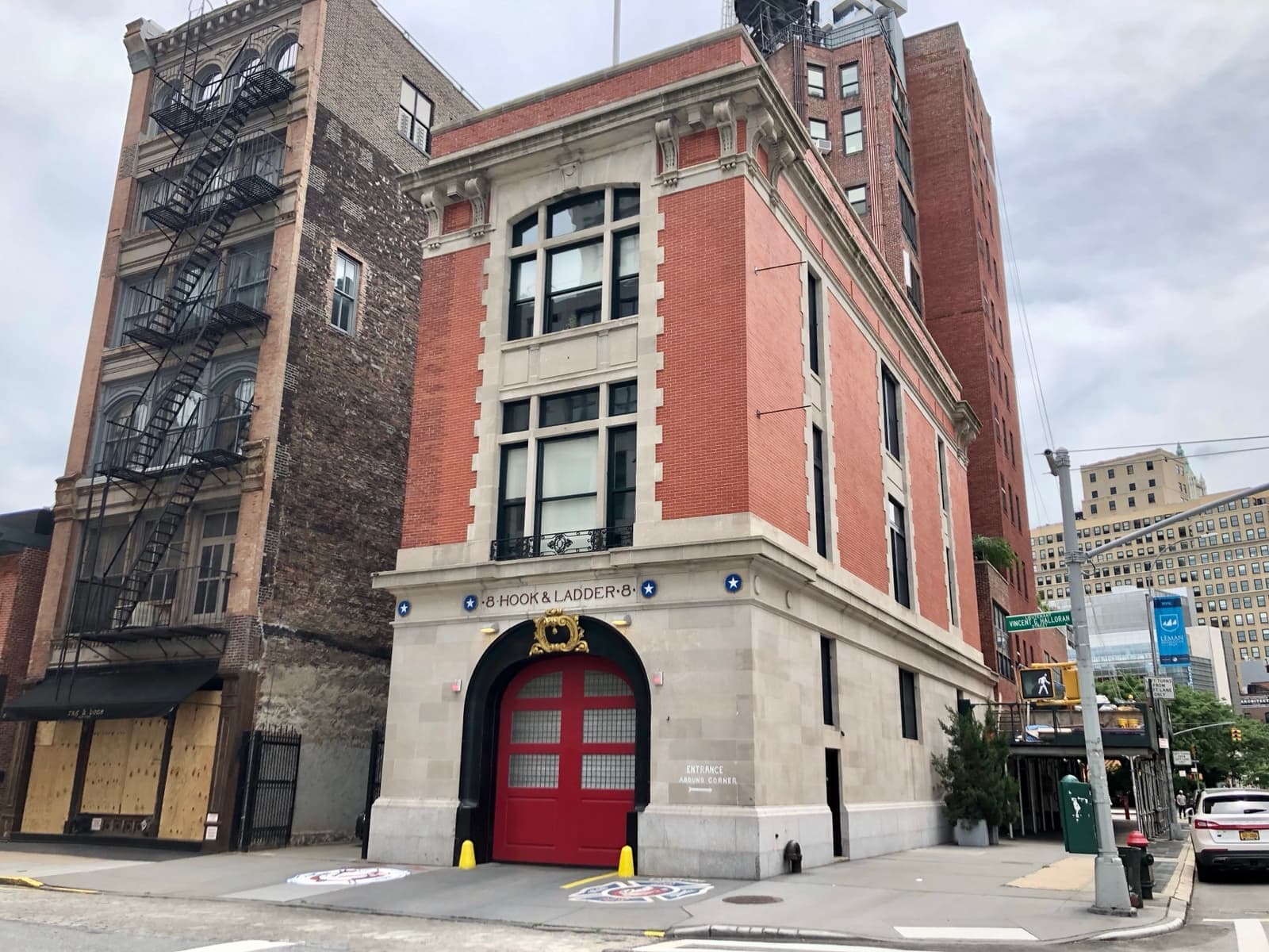 The firehouse on its corner beside a taller fire-escaped building, red doors shut