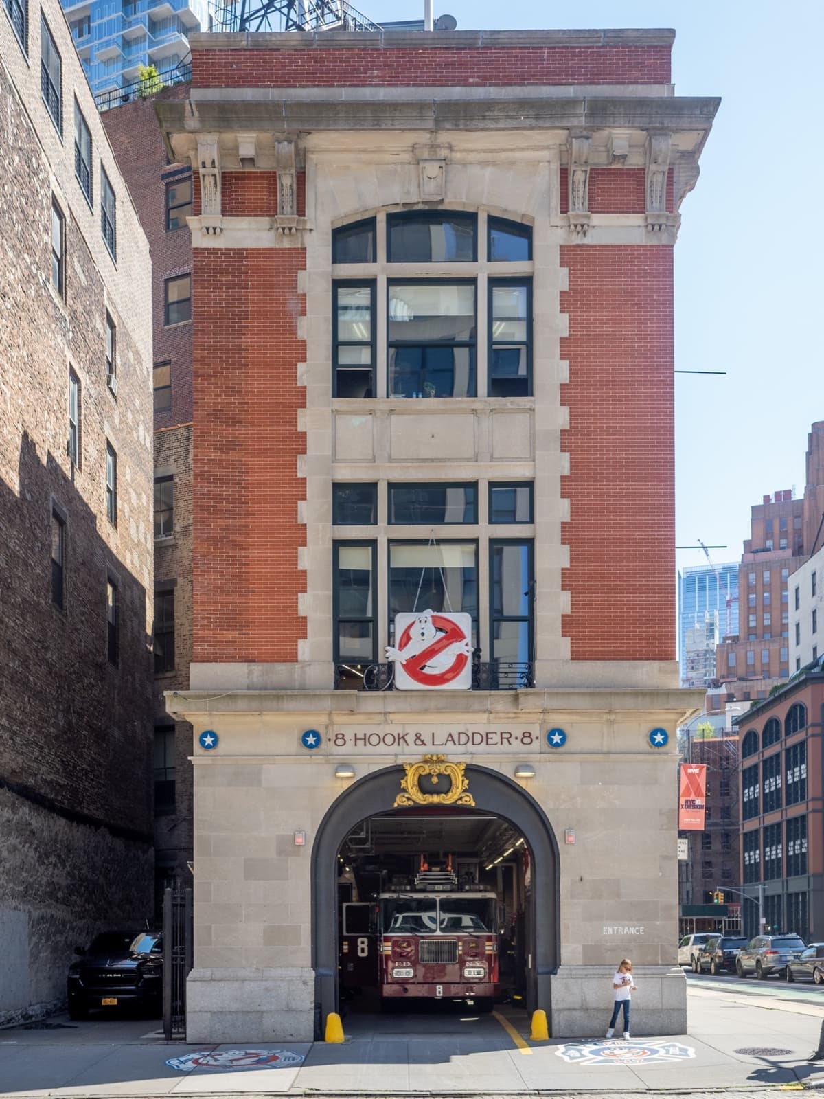 A narrow red-brick and limestone firehouse with its bay door open and a ladder truck inside
