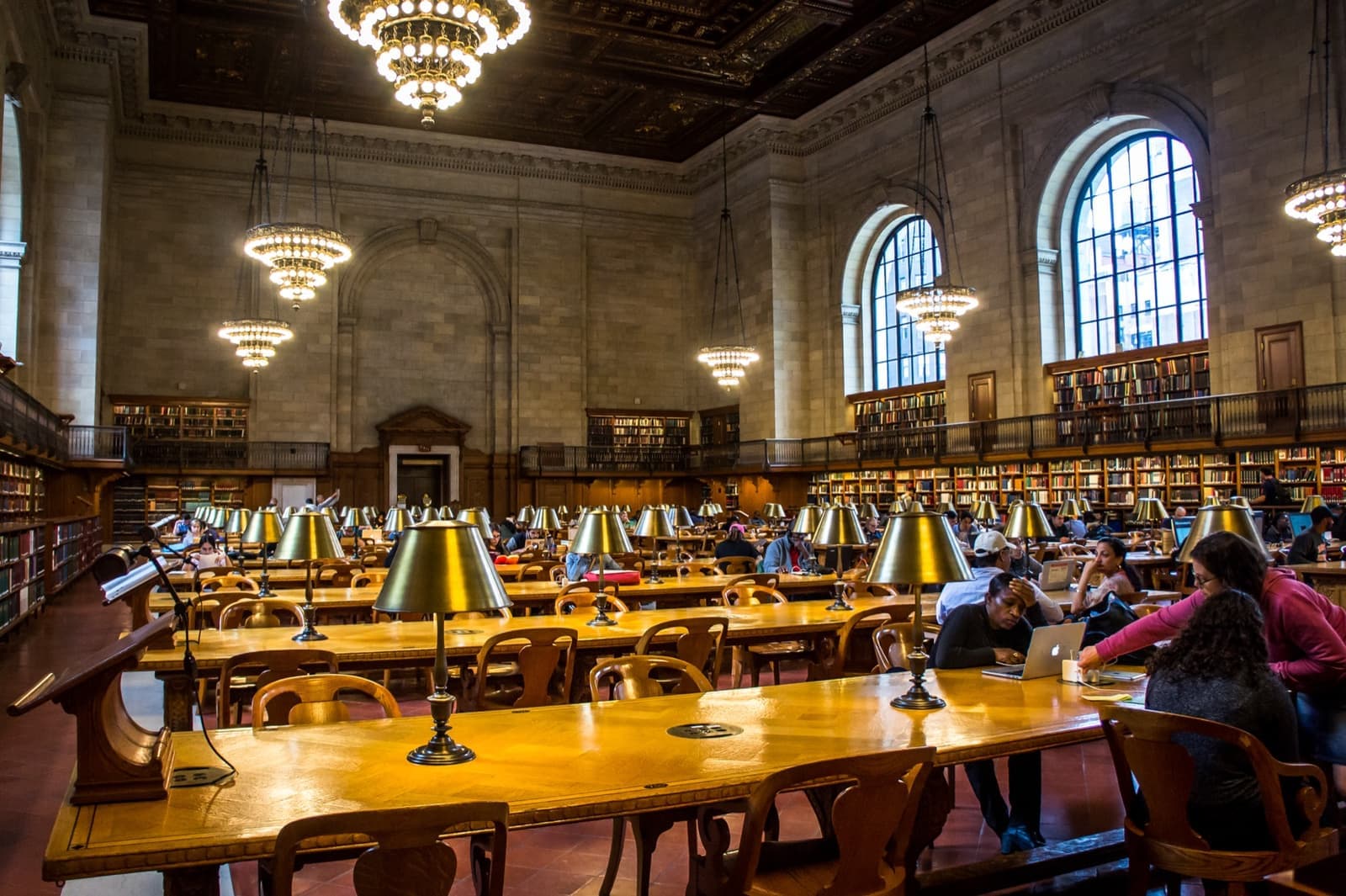 A long reading room with chandeliers, arched windows and rows of lamps on wooden tables