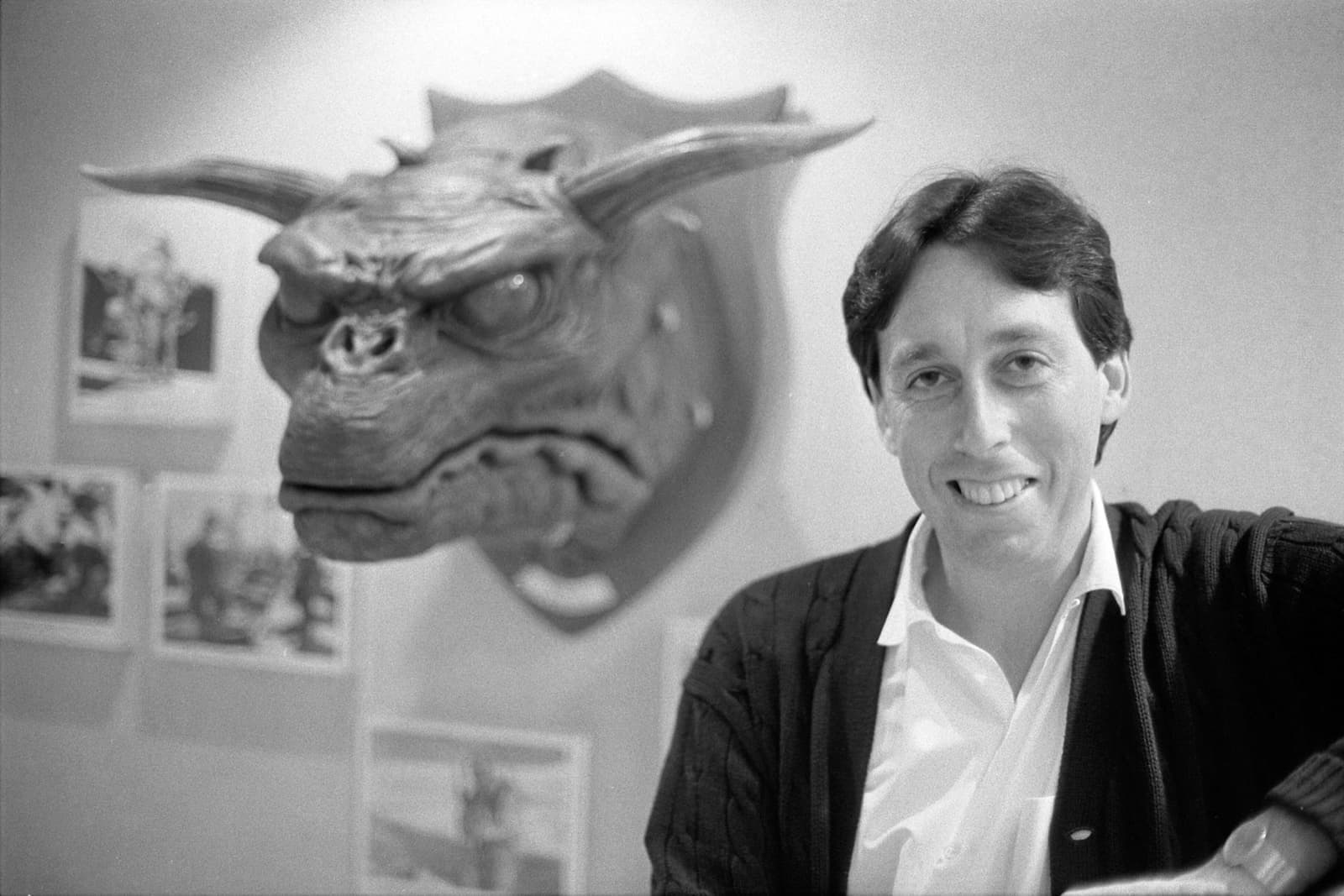 Ivan Reitman, smiling in a dark cardigan, beside a horned creature head mounted on a wall