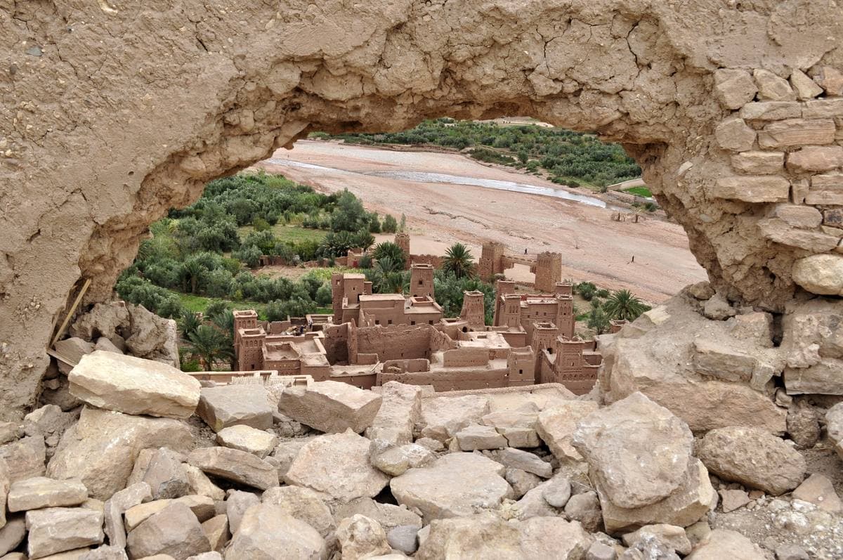 Earthen houses seen through a ragged stone gap, with green palms and a pale riverbed beyond