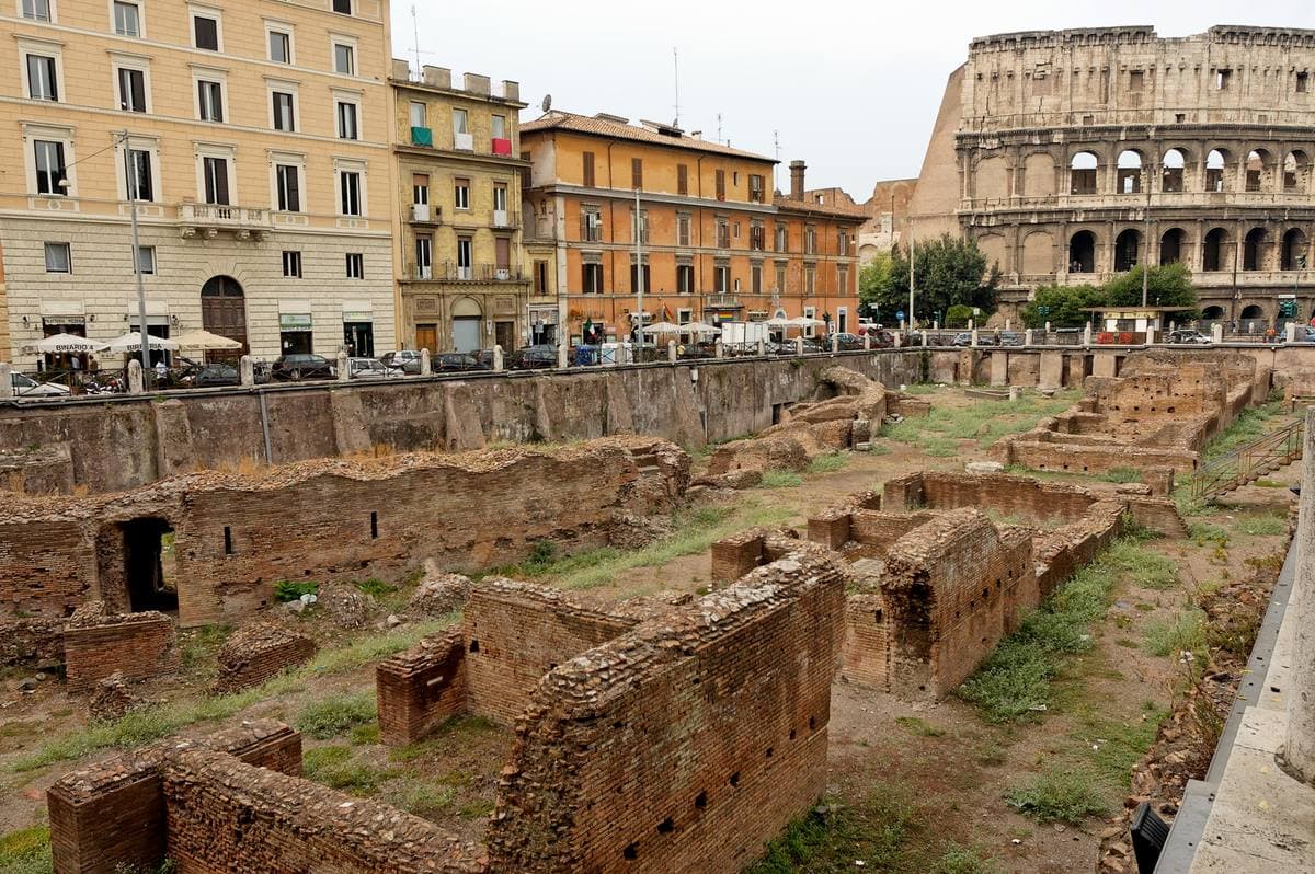 Low brick walls of an excavated ruin below a street, the Colosseum at the end of the road