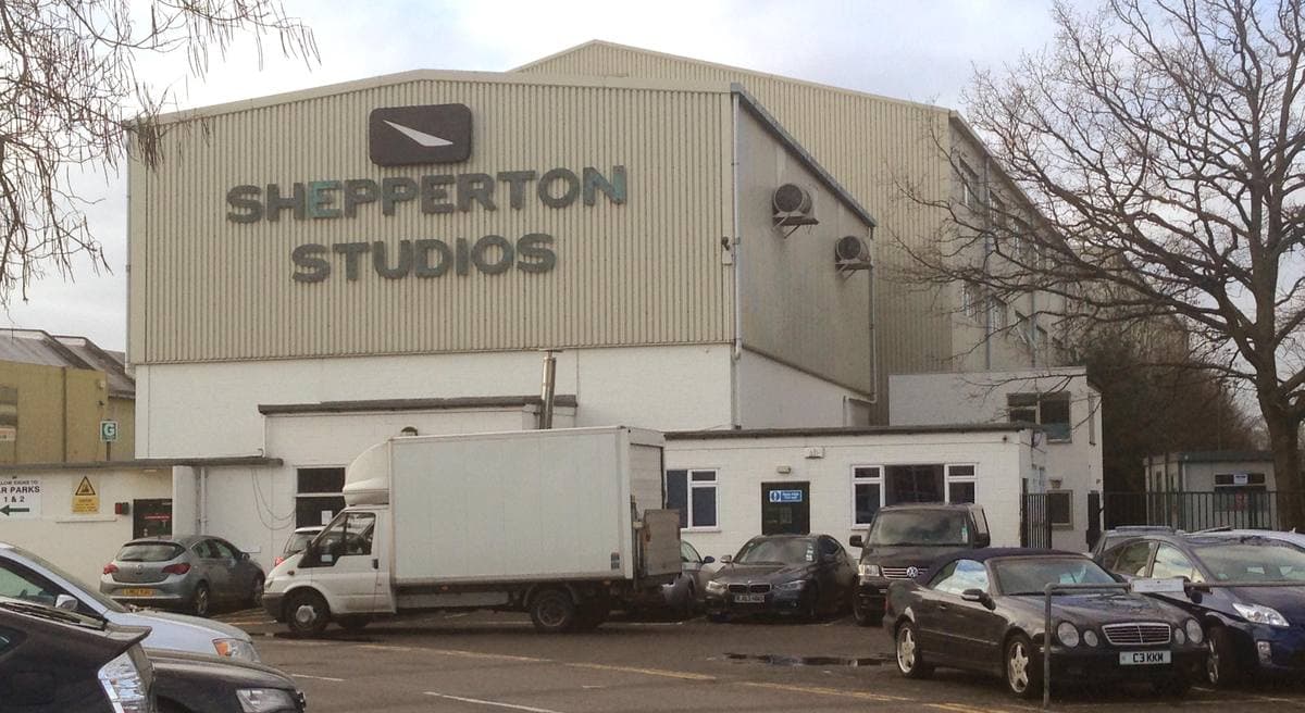 A grey studio building with a Shepperton Studios sign over parked cars