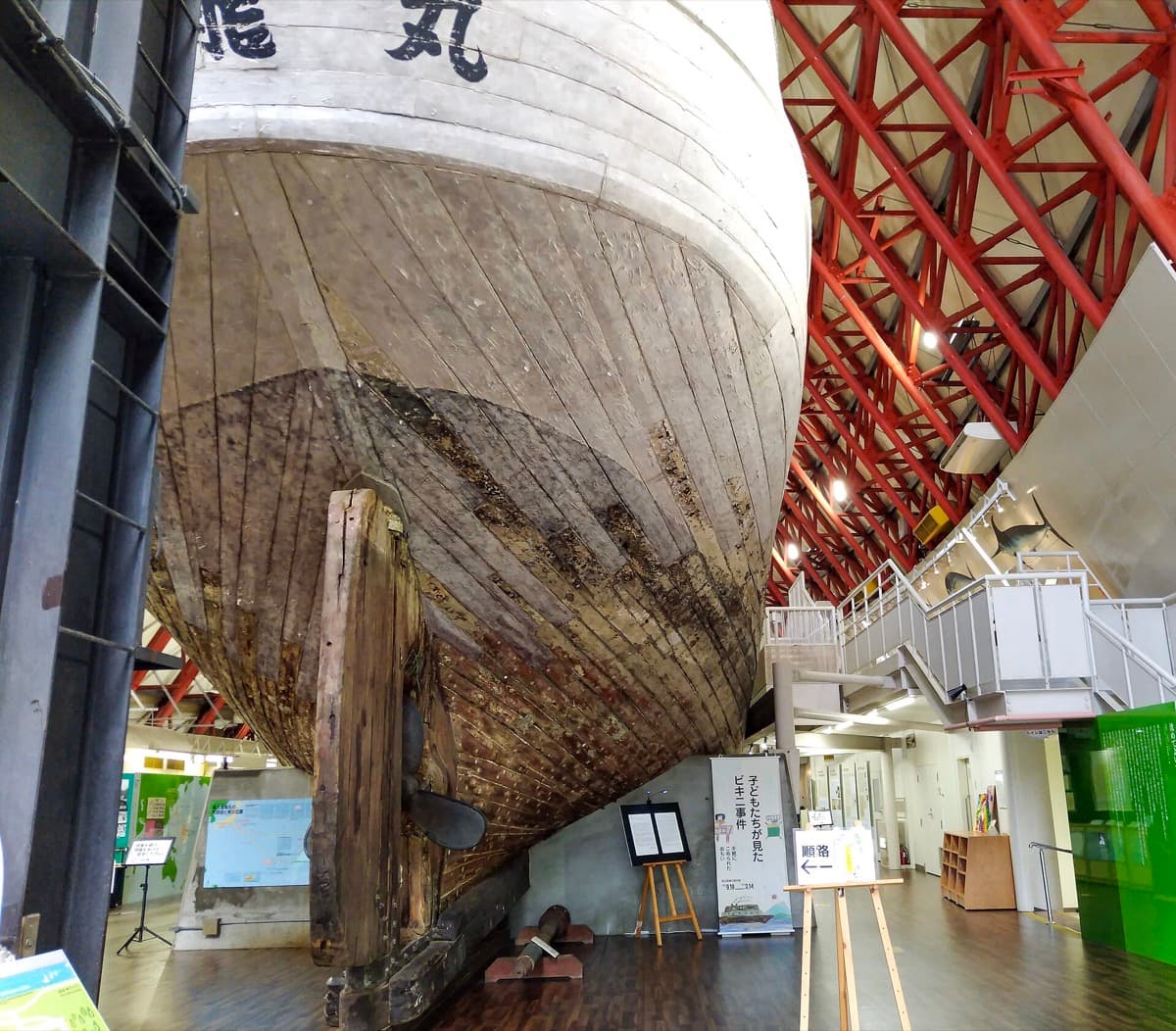 The wooden hull of a fishing boat raised under a red steel roof inside a museum hall
