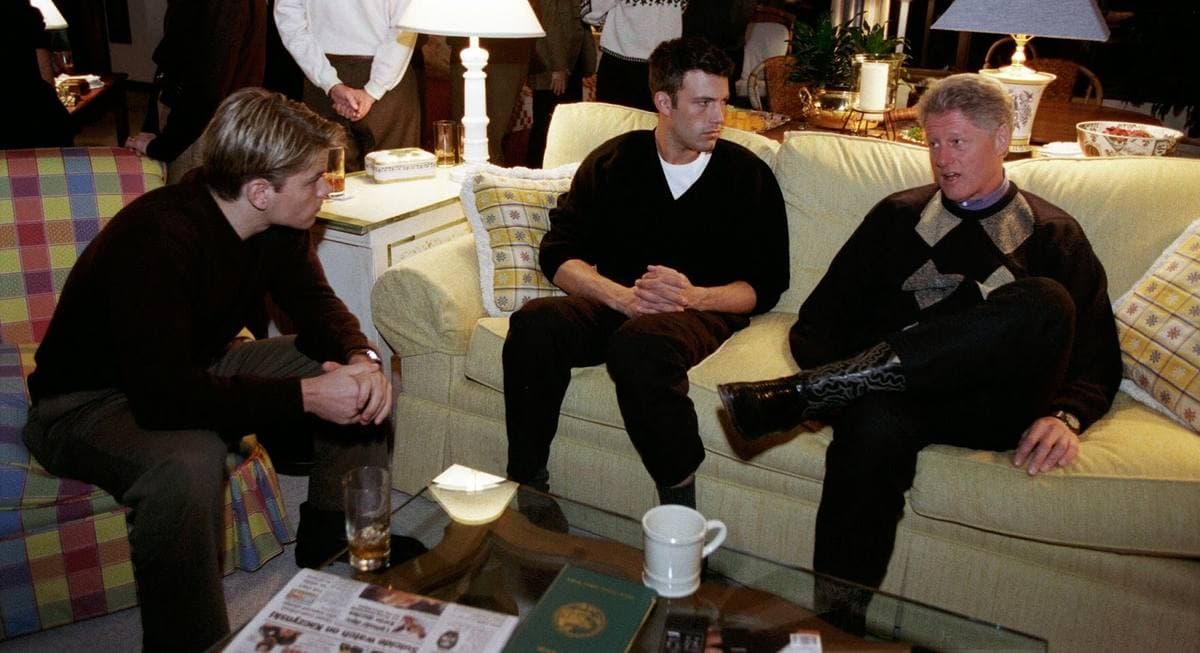Matt Damon, Ben Affleck and Bill Clinton sitting on sofas in a lamp-lit lounge, talking