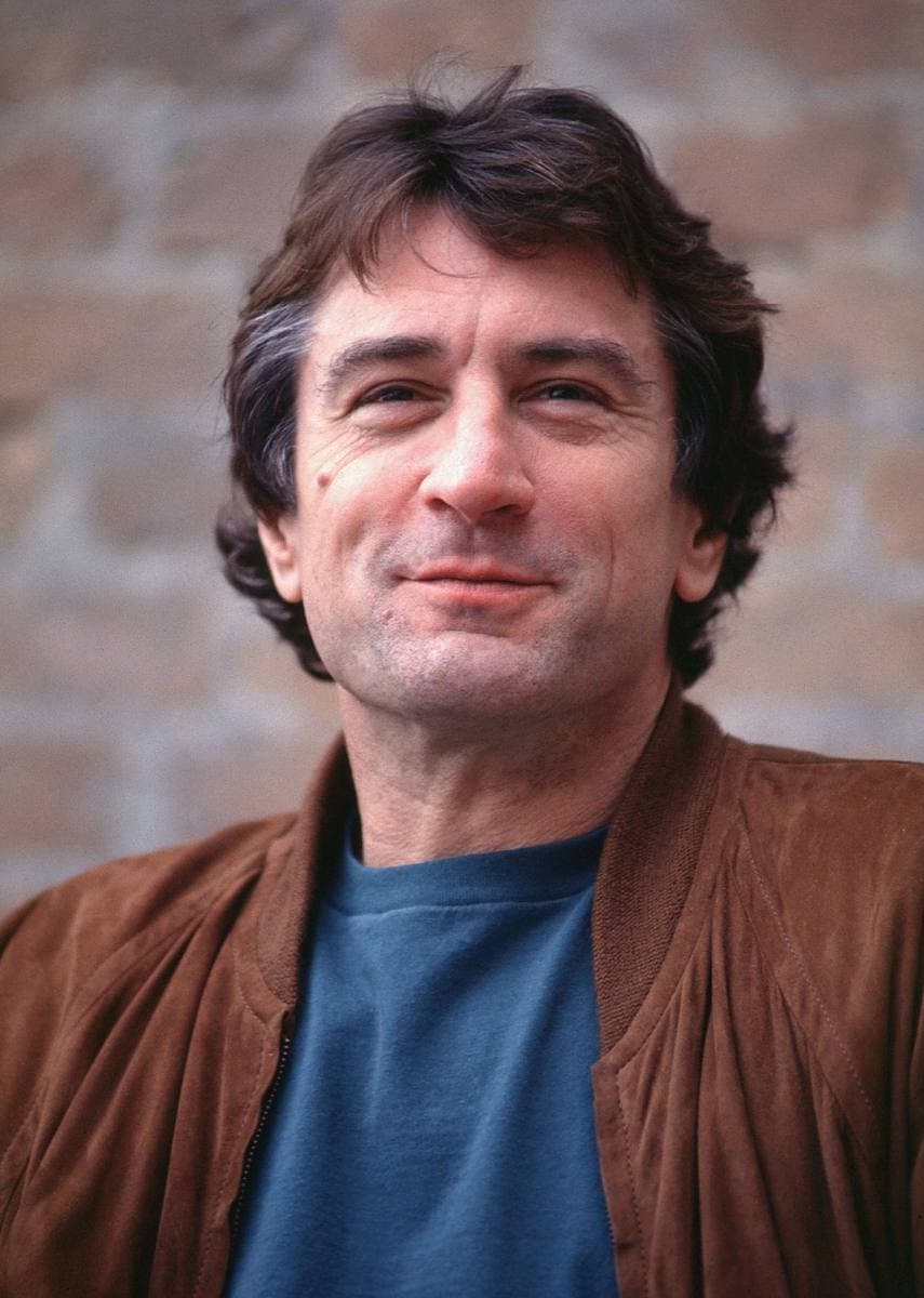 Colour photograph of Robert De Niro in a brown leather jacket, smiling against a brick wall