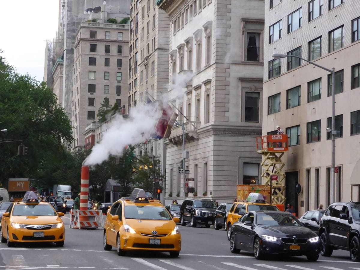 Colour photograph of yellow taxis on Fifth Avenue by stone apartment buildings, steam rising from a street pipe
