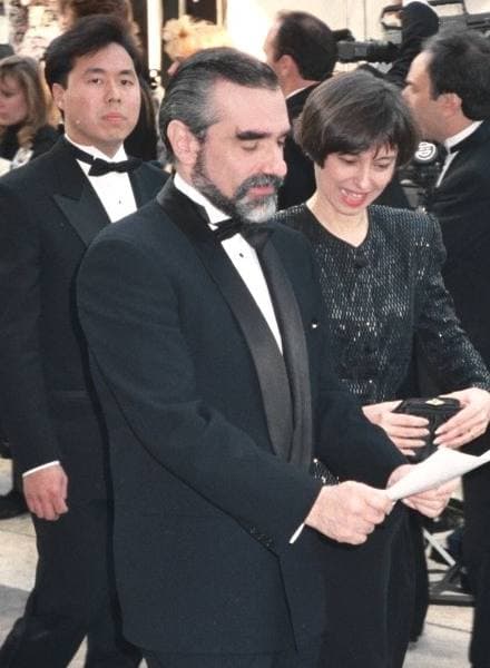Martin Scorsese in a dinner jacket and bow tie, bearded, reading a card on the red carpet